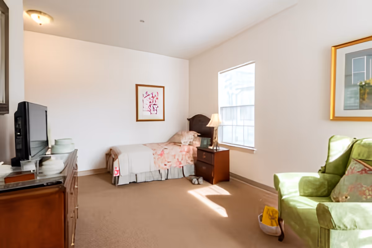 A bright bedroom with a single bed covered in a floral bedspread, a wooden nightstand with a lamp, a green upholstered armchair, a dresser with a TV on top, and two framed pictures on the walls. Sunlight streams in through a large window.