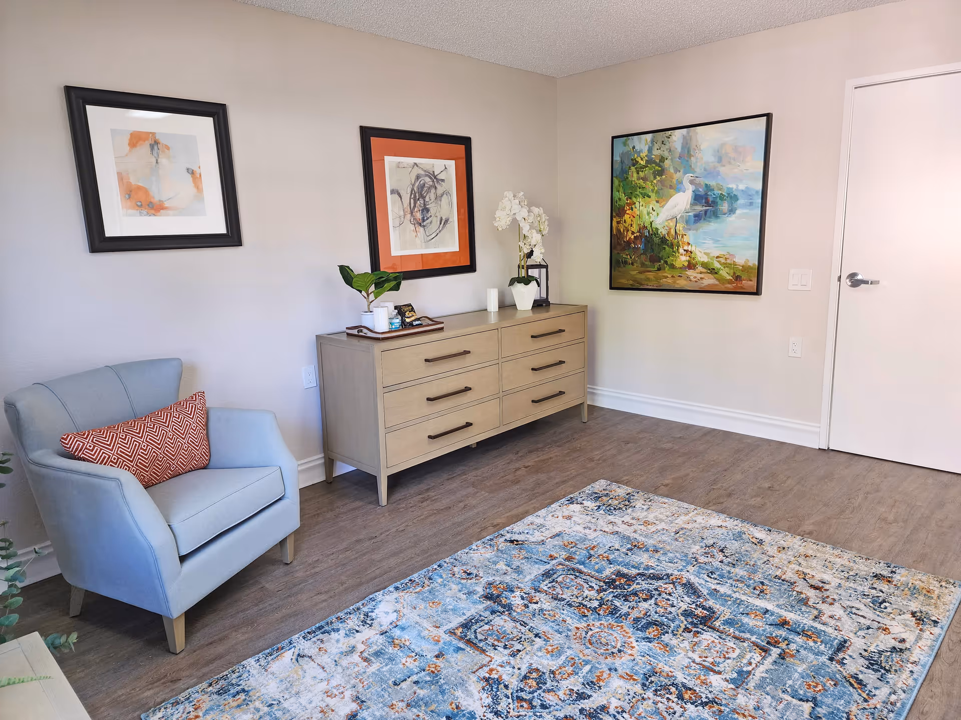 A cozy interior room featuring a light blue armchair with a red patterned pillow, a wooden dresser with six drawers adorned with a potted plant, a white orchid, and decorative items. The walls are decorated with three framed artworks, and a colorful patterned rug covers part of the wooden floor. A closed white door is visible on the right side.