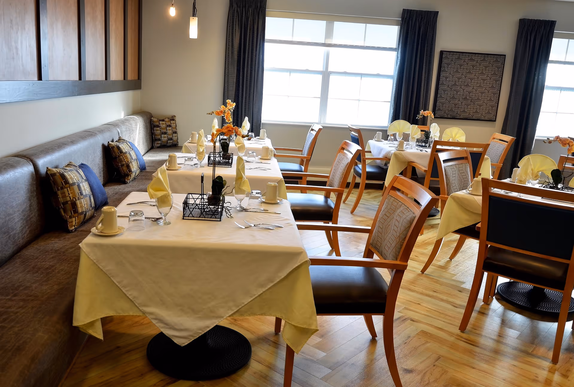 A bright dining room with several tables covered in white tablecloths and set with cups, glasses, napkins, and silverware. The tables are surrounded by wooden chairs with cushioned seats and backs. A cushioned bench with patterned pillows runs along one wall. Large windows with dark curtains allow natural light to fill the room. Decorative flowers and framed artwork add to the cozy atmosphere.