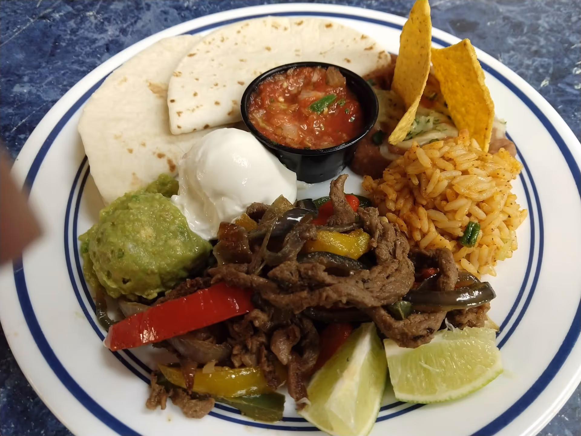Plate of Mexican-style food with sliced beef and peppers, rice, guacamole, sour cream, salsa, tortillas, chips, and lime wedges.