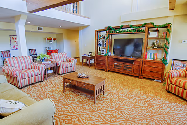 A spacious living room with patterned carpet, a large wooden entertainment center with a flat-screen TV, and several striped armchairs and a beige sofa arranged around a wooden coffee table. The room has high ceilings with exposed wooden beams and a balcony railing visible above.