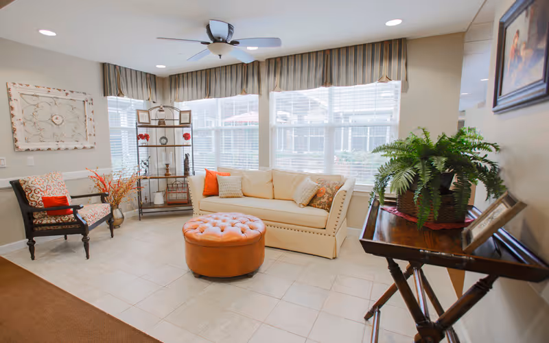 Bright lounge area with a beige sofa, patterned armchair, round leather ottoman, and large windows.
