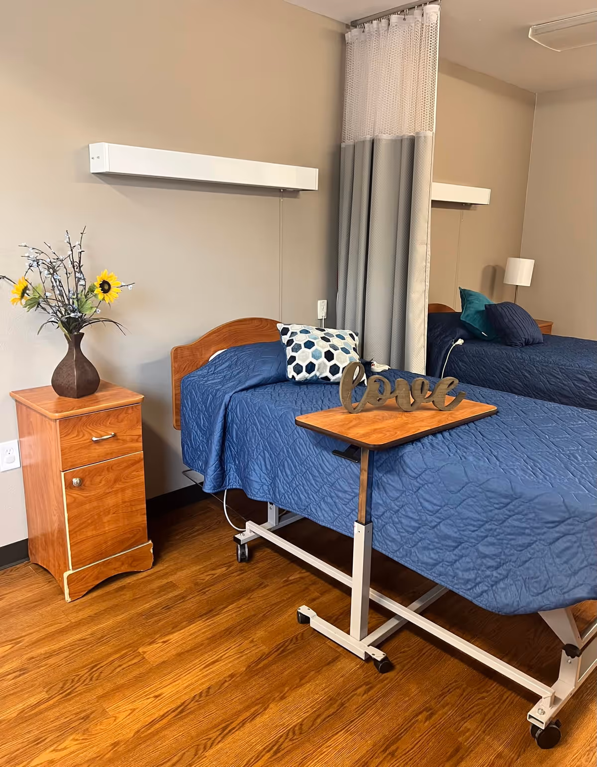 A nursing facility room with two hospital beds covered in blue quilts, separated by a privacy curtain. One bed has a decorative pillow and a wooden tray table with a decorative 'love' sign. There is a wooden nightstand with a vase of sunflowers and other flowers on it. The room has wooden flooring and beige walls.