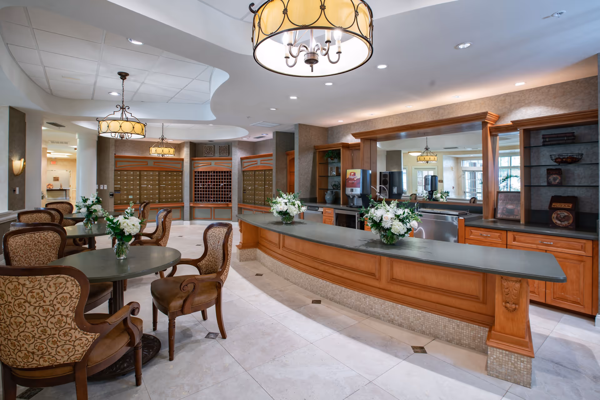 A spacious and elegant common area in a senior living facility featuring a long wooden counter with a green countertop, decorated with white floral arrangements. There are several round tables with upholstered chairs arranged for seating. The room has soft lighting from hanging chandeliers and wall sconces. In the background, there are mailboxes and a decorative wooden shelving unit.