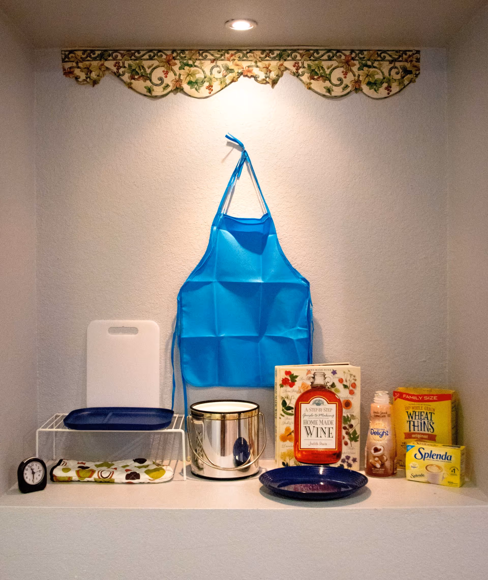 A kitchen nook with a blue apron hanging on the wall, a white cutting board, a blue plate, a metal container, a cookbook titled 'Home Made Wine', a bottle of coffee creamer, a box of Wheat Thins crackers, a box of Splenda sweetener, a small clock, and a folded kitchen towel with fruit patterns.