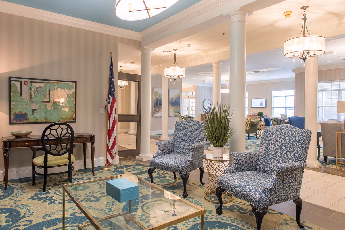 A bright and spacious senior living facility common area with patterned blue armchairs, a glass coffee table, a small side table with a potted plant, and a wooden desk with a chair. The room features white columns, hanging light fixtures, an American flag, and framed artwork on the walls. Large windows allow natural light to fill the space.