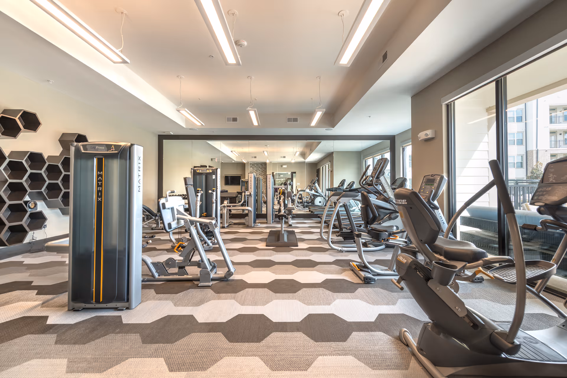 A modern fitness room with various exercise equipment including stationary bikes, leg press machines, and weight machines. The room has large windows letting in natural light, a patterned carpet floor, and a large mirror covering one wall.