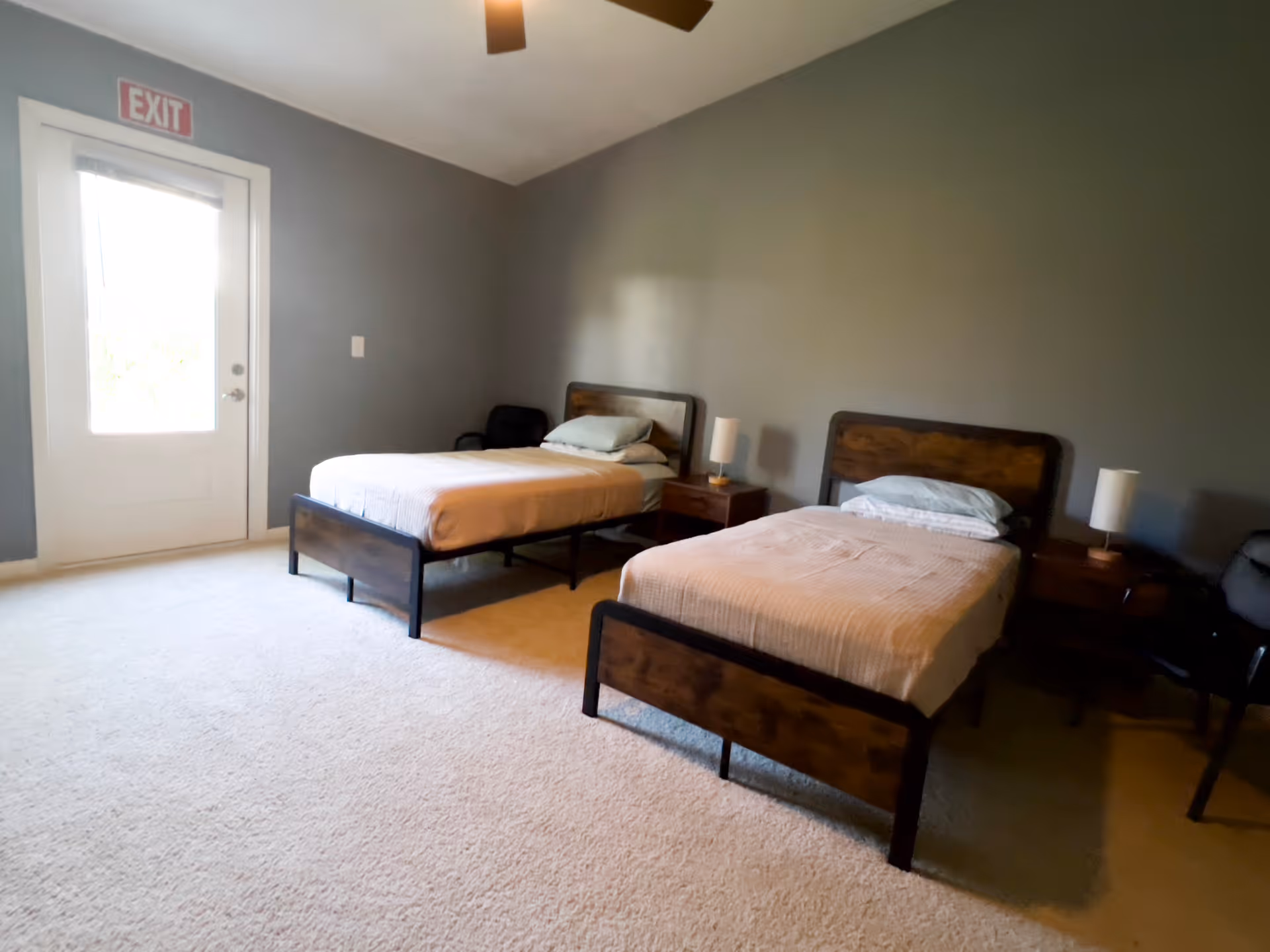 A bedroom with two single beds, each with a wooden headboard and footboard, white bedding, and a pillow. Between the beds is a wooden nightstand with a small lamp. There are two chairs, one near each bed. The walls are painted gray, and there is a door with an exit sign above it. The floor is carpeted in a light color.
