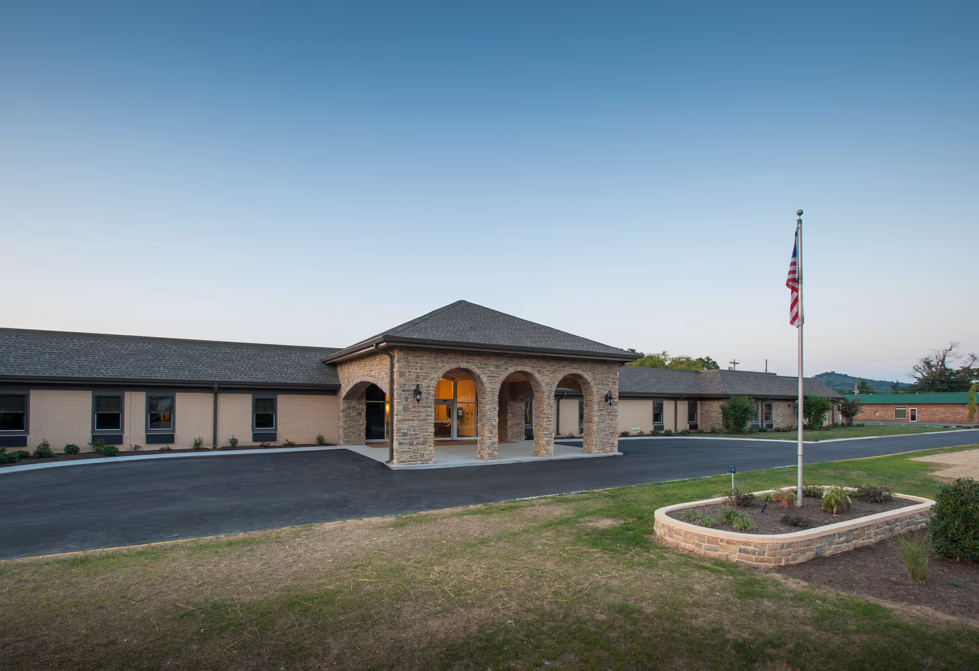 Single-story brick senior living facility front entrance with a covered porte-cochère and an American flag on a flagpole.