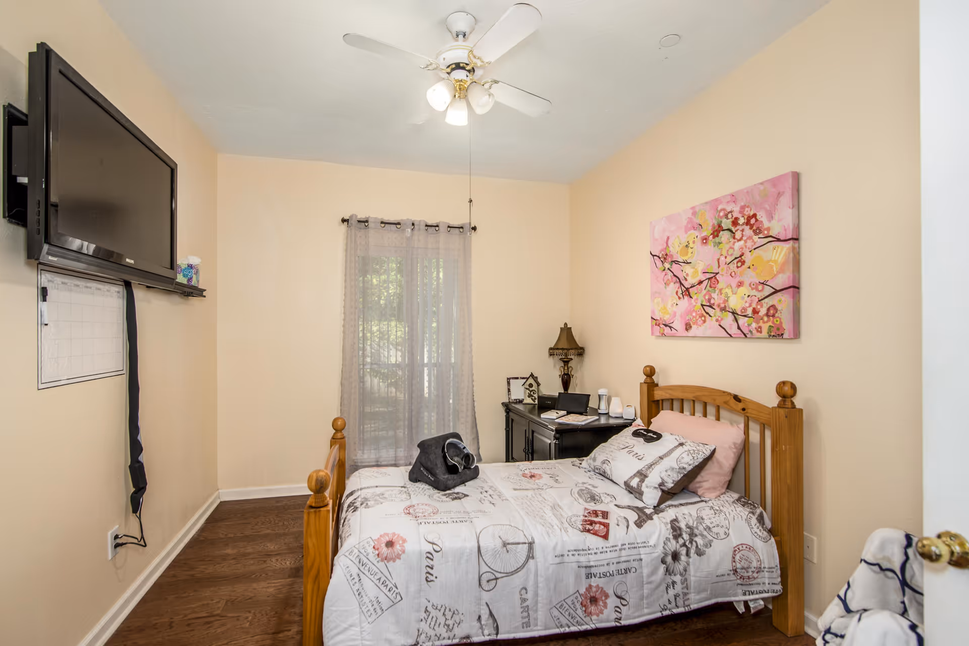 A small bedroom with a wooden single bed covered with a white and pink patterned quilt and pillows. There is a black desk with a lamp, a laptop, and some items on it against the wall. A sheer curtain covers a window behind the desk. A flat-screen TV is mounted on the wall opposite the bed. A ceiling fan with lights is on the ceiling, and a pink floral painting hangs above the bed.