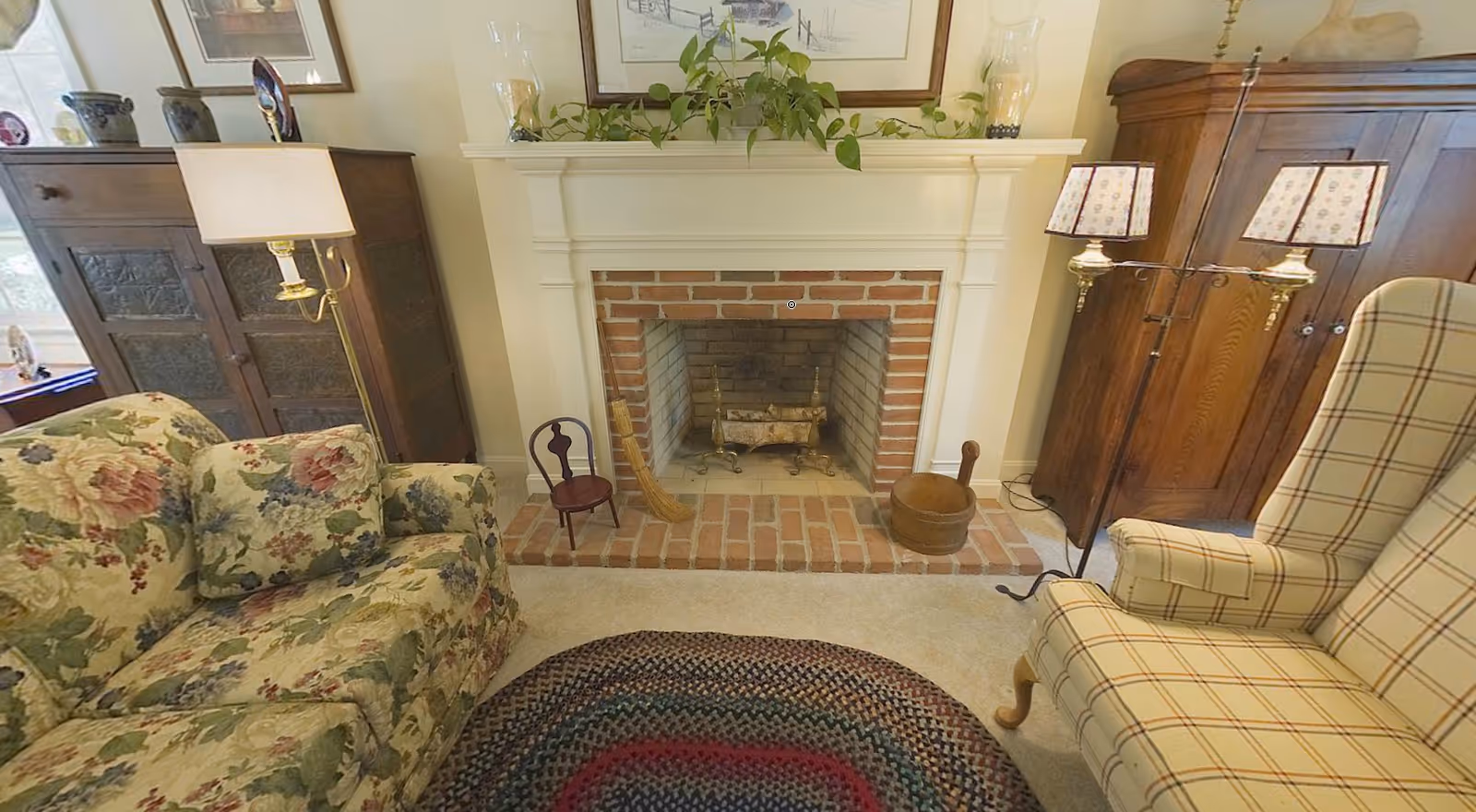 Cozy living room with a brick fireplace in the center, flanked by a floral patterned sofa on the left and a plaid upholstered armchair on the right. The fireplace mantel is decorated with green plants and framed artwork above. There are wooden cabinets on both sides of the fireplace, a floor lamp with a white shade next to the sofa, and a double-shaded lamp near the armchair. A colorful braided rug lies on the carpeted floor in front of the fireplace.