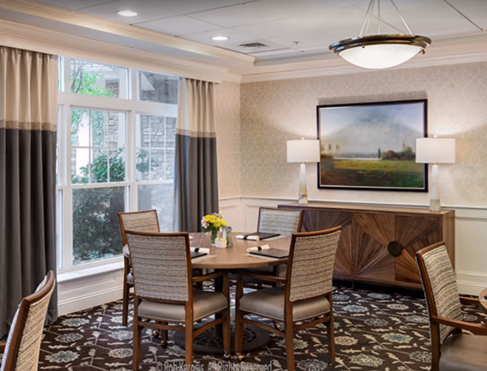 A dining room with a round wooden table surrounded by six upholstered chairs. The room has a large window with beige and dark gray curtains, a patterned carpet, and a wooden sideboard with two table lamps and a landscape painting above it. A ceiling light fixture hangs above the table.