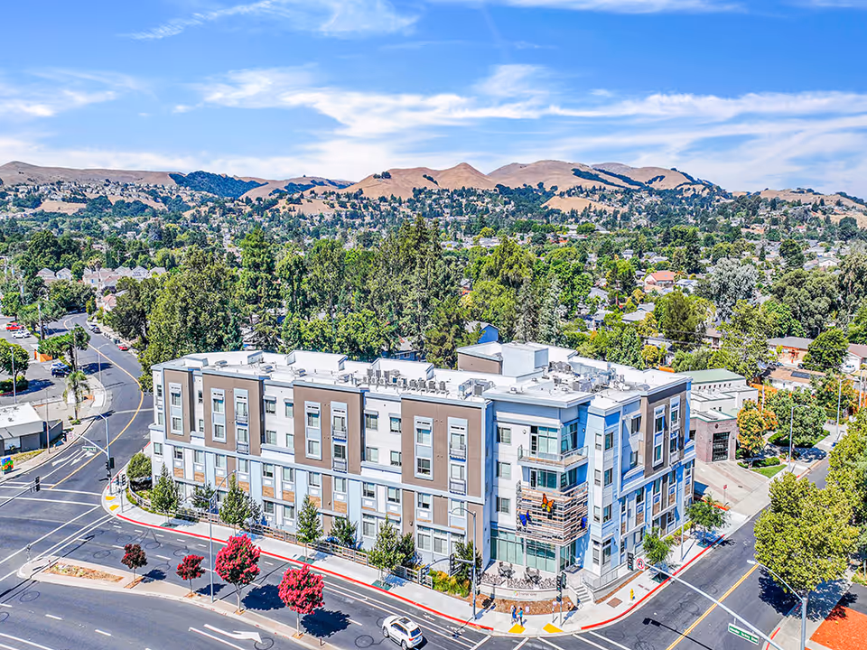 Aerial view of a modern, multi-story senior living facility named Emerald Care Home located at a street corner with surrounding trees, roads, and a suburban neighborhood with hills in the background under a partly cloudy sky.