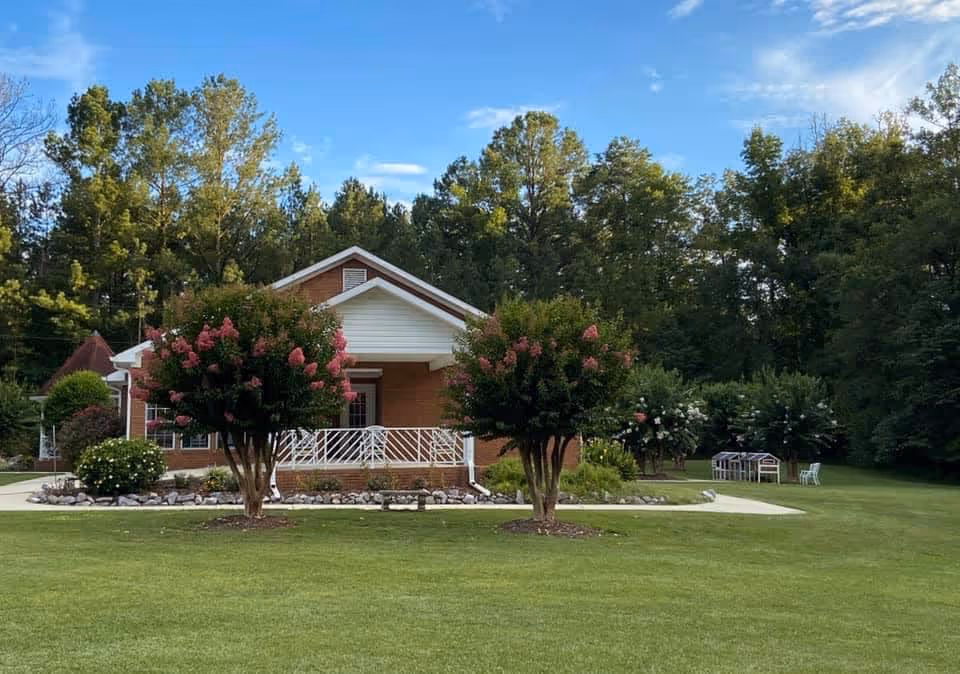 A single-story brick building with a white porch railing, surrounded by green grass and trees. Two flowering trees with pink blossoms are in front of the building, and there is a small white structure and chairs on the lawn to the right. The sky is blue with some clouds.