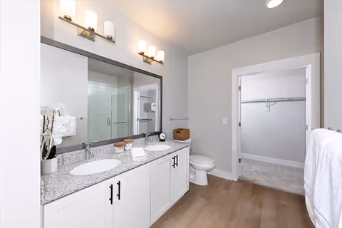 A modern bathroom with a double sink vanity featuring a granite countertop, two mirrors with light fixtures above, a toilet, and a walk-in closet visible through an open door. The bathroom has light-colored walls and wood-style flooring.