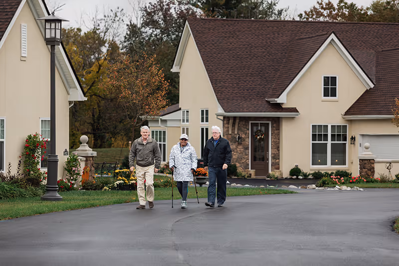 Three elderly people walking on a paved road in front of beige houses with brown roofs. One person is using a cane and another is using a walker. The scene is outdoors with trees and greenery in the background.