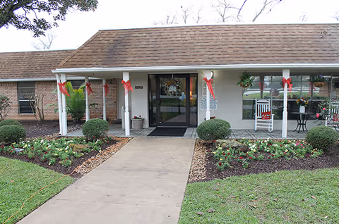 Front entrance of Creekside Village Healthcare facility with a covered porch decorated with red bows and hanging plants. There are two white rocking chairs and a small table on the porch. The walkway is flanked by neatly maintained flower beds and bushes.