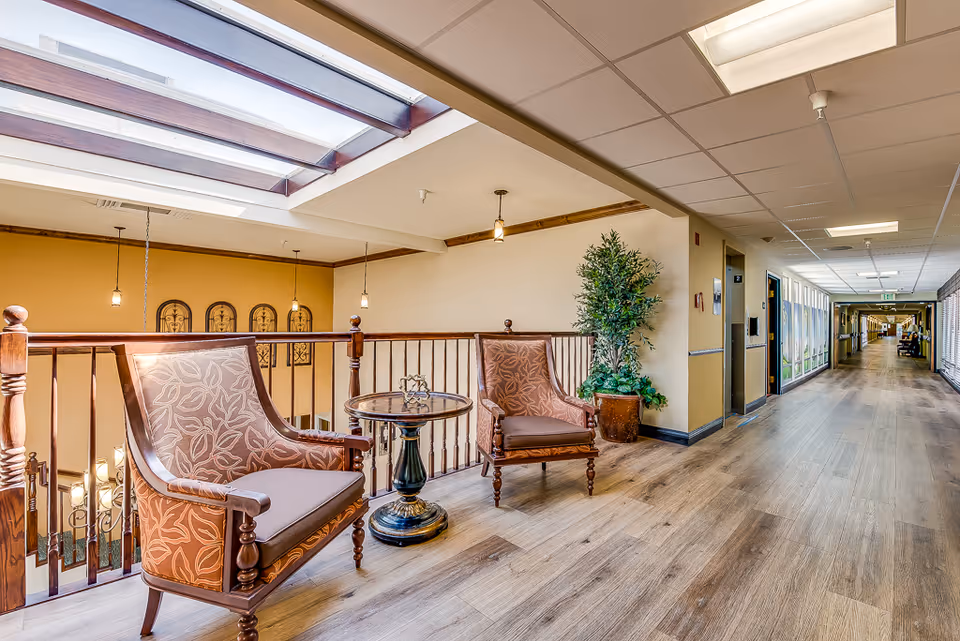 Upper-level interior hallway with two upholstered chairs and a small table beside a railing and potted plant under skylights.