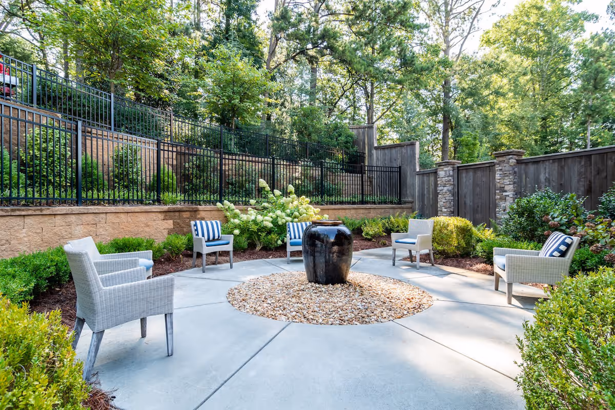 Outdoor seating area with five wicker chairs arranged in a circle around a central black water fountain on a bed of small rocks. The area is surrounded by greenery, bushes, and trees, with a stone and wooden fence enclosing the space.
