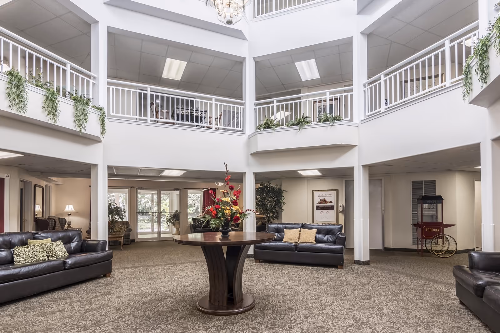 Spacious two-story common area in a senior living facility with leather sofas arranged around a central round wooden table adorned with a floral arrangement. The upper level features white railings with hanging green plants, and the area is well-lit with ceiling lights. A popcorn machine is visible in the corner near the wall.