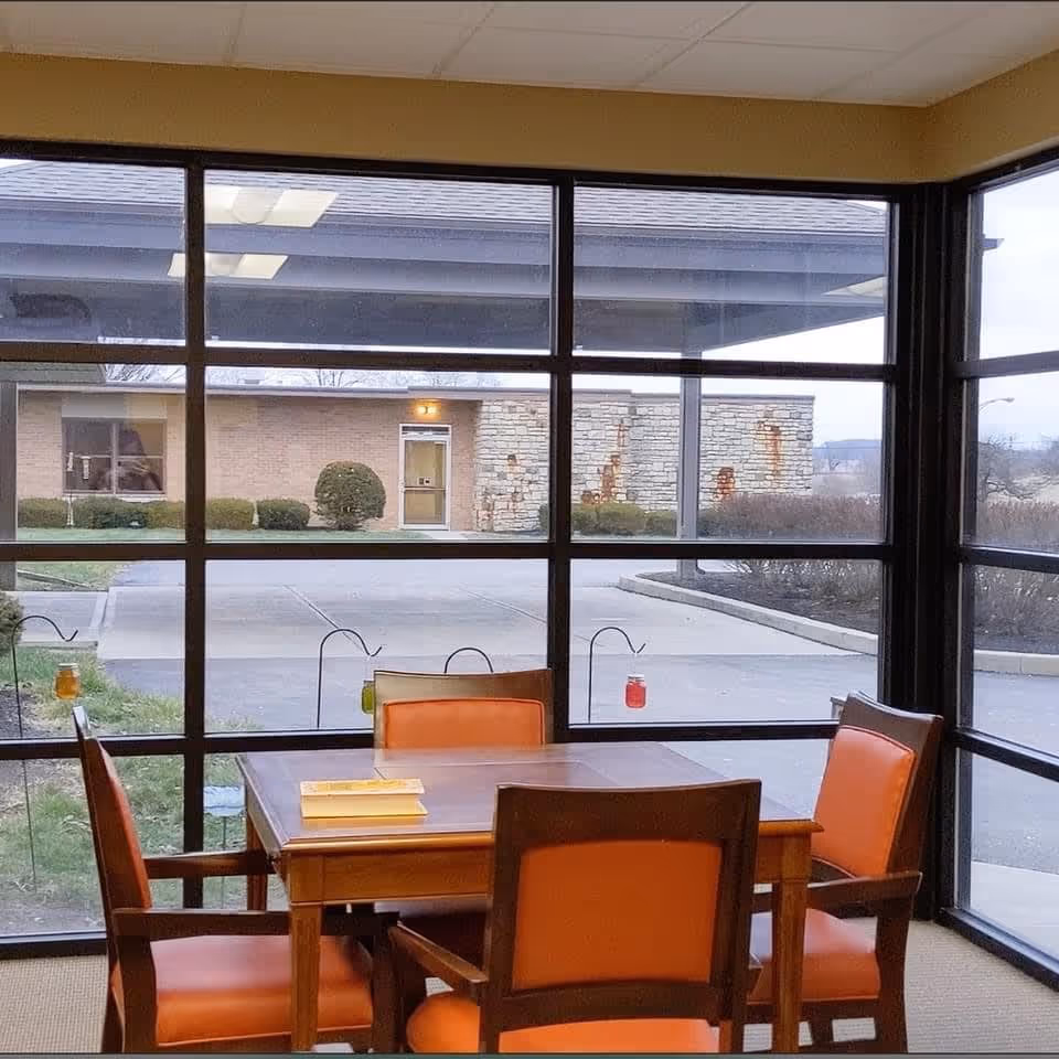 Interior view of a room with a wooden table and orange chairs, looking out through large windows at the entrance of Vancrest of Urbana, featuring a landscaped area and a brick building.