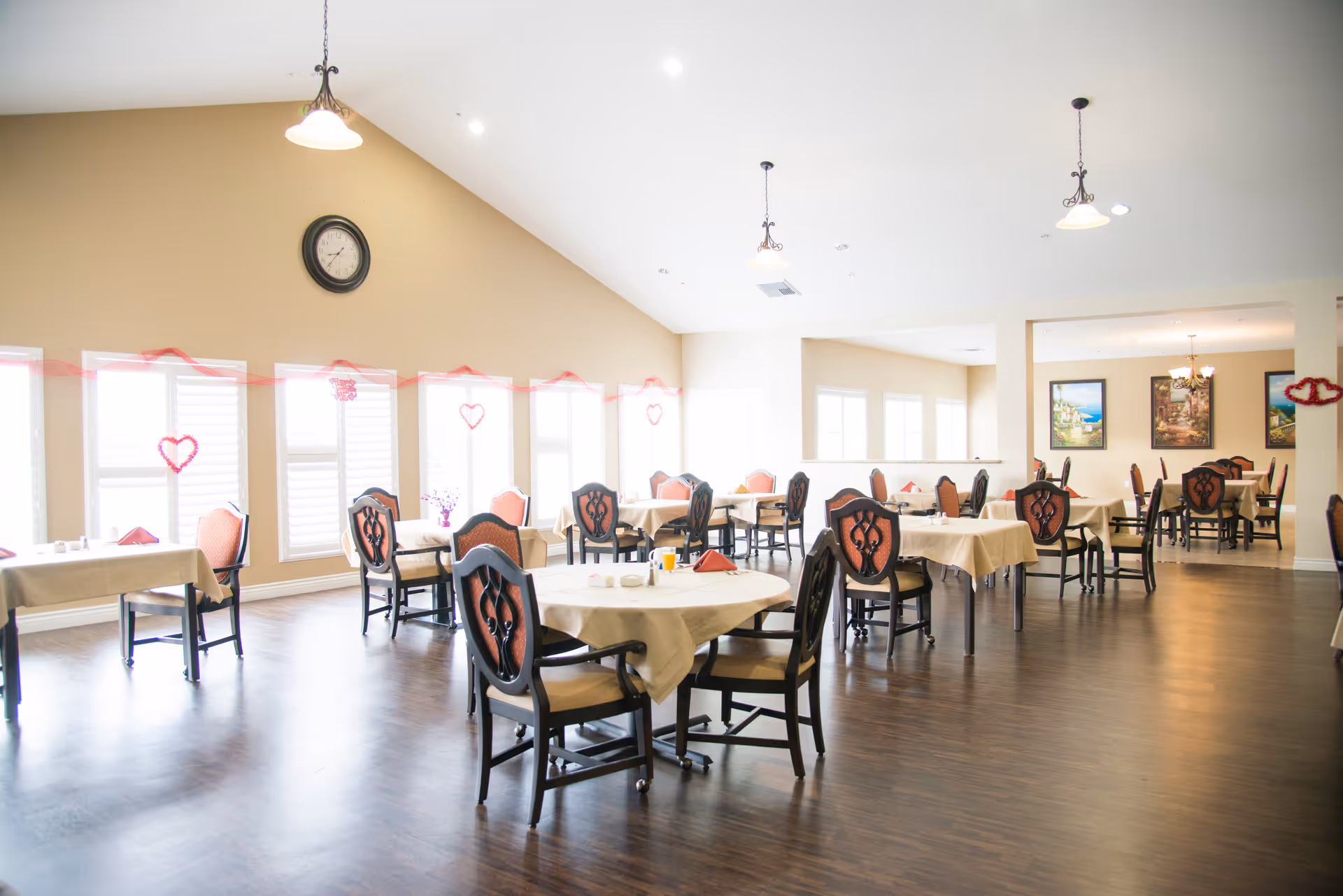 A spacious dining room with multiple tables covered in beige tablecloths and surrounded by chairs with red cushioned backs. The room has large windows with white shutters letting in natural light, a high vaulted ceiling with hanging light fixtures, and a clock on the beige wall. Red heart-shaped decorations are hung along the windows.