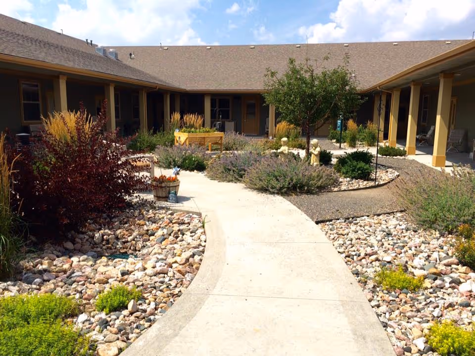 Outdoor courtyard area of Mountain Plaza Assisted Living & Memory Care featuring a curved concrete walkway surrounded by decorative rocks, various shrubs, plants, and small trees. The courtyard is enclosed by a single-story building with a covered walkway supported by columns.