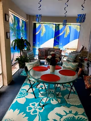 Covered sunroom-style seating area with a round glass table with red placemats on a turquoise floral rug, surrounded by cushioned chairs, a sofa, tropical curtains, and plants.