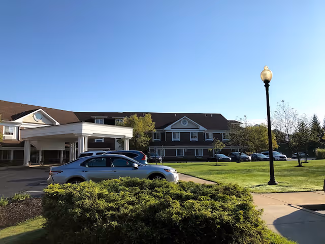 Exterior view of a senior living facility named Parkside Villa with a parking area in front, several cars parked, a well-maintained lawn, bushes, trees, and a street lamp under a clear blue sky.