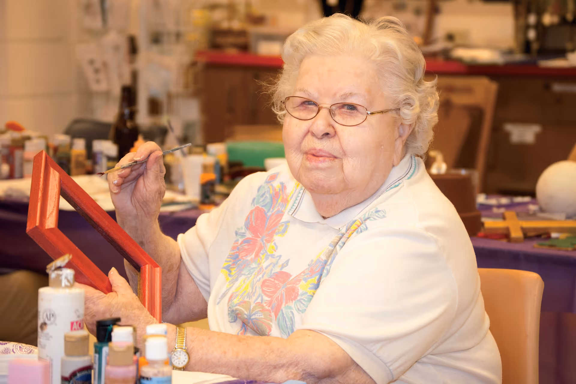 An elderly woman with white hair and glasses is sitting at a table engaged in painting a red picture frame. Various paint bottles and art supplies are visible on the table around her, and the background shows a room with more art materials and furniture.