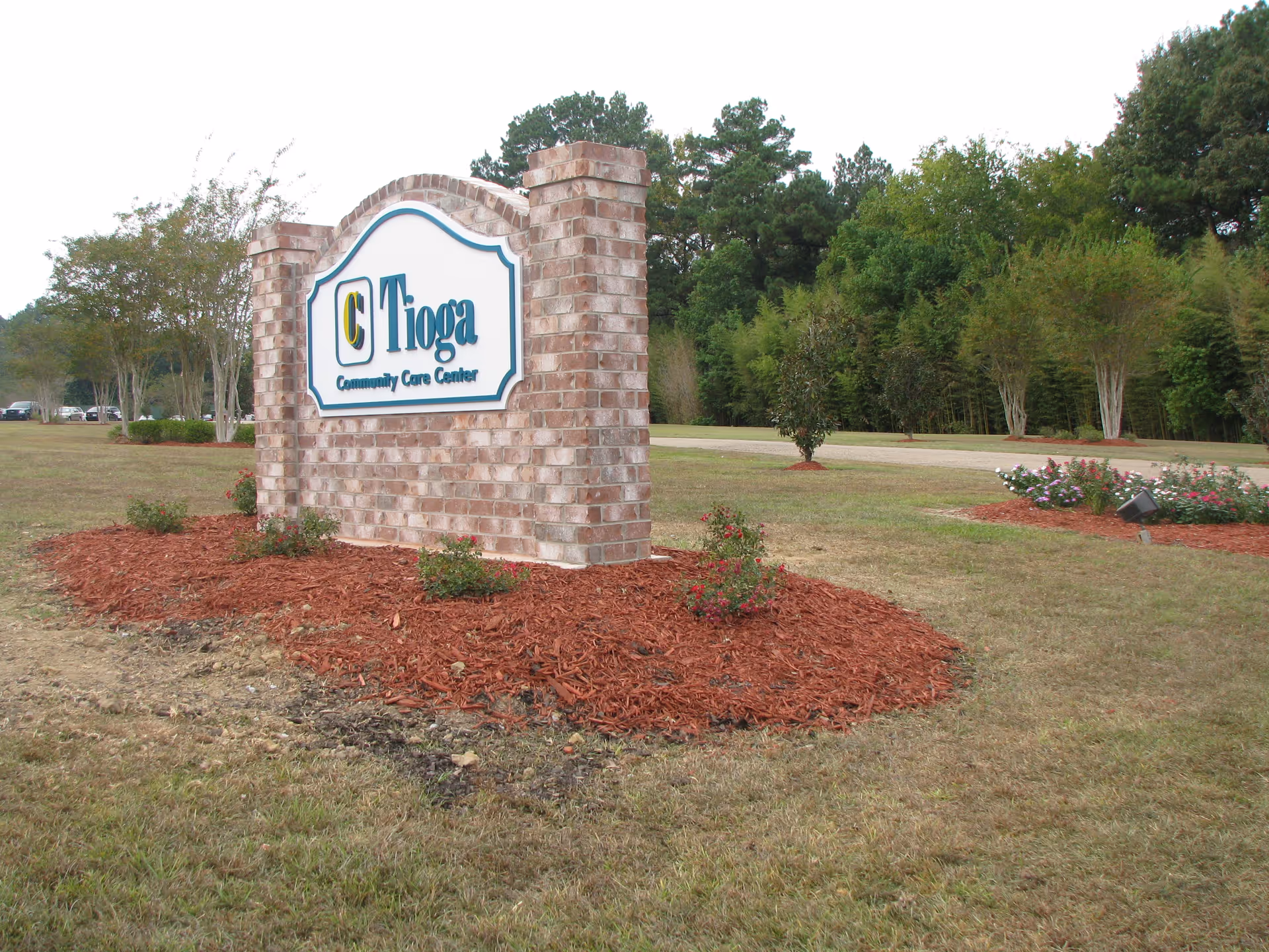 A brick sign for Tioga Community Care Center surrounded by mulch and small bushes, with trees and a grassy area in the background.