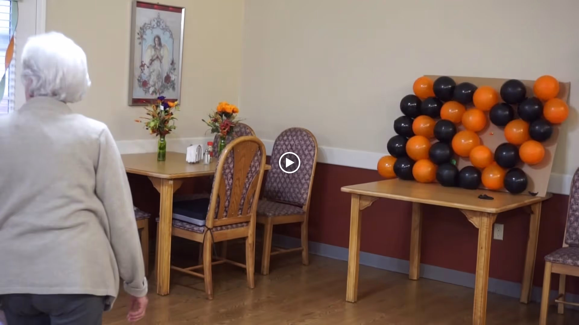 An elderly woman walks past a dining area with wooden tables and chairs, floral centerpieces, and a board of orange and black balloons.
