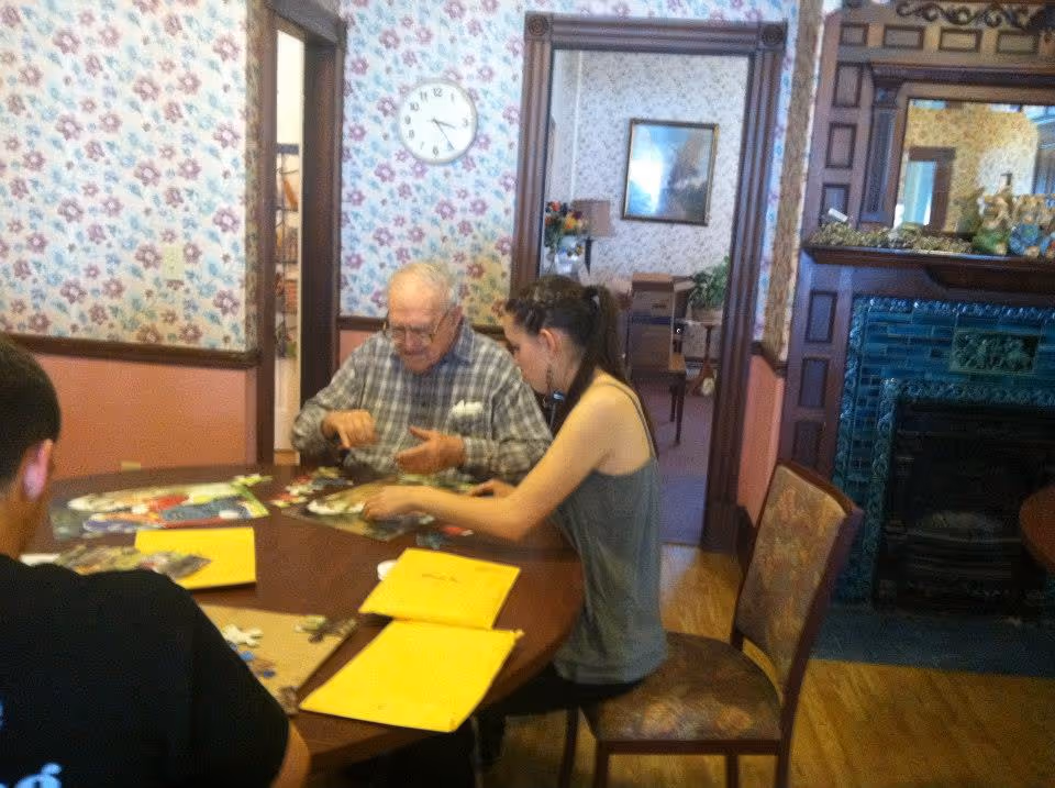 An elderly man and a young woman sitting at a round wooden table working on a puzzle together in a room with floral wallpaper and a wooden fireplace with green tiles. There is a clock on the wall showing the time as 3:40, and some yellow folders or papers on the table.