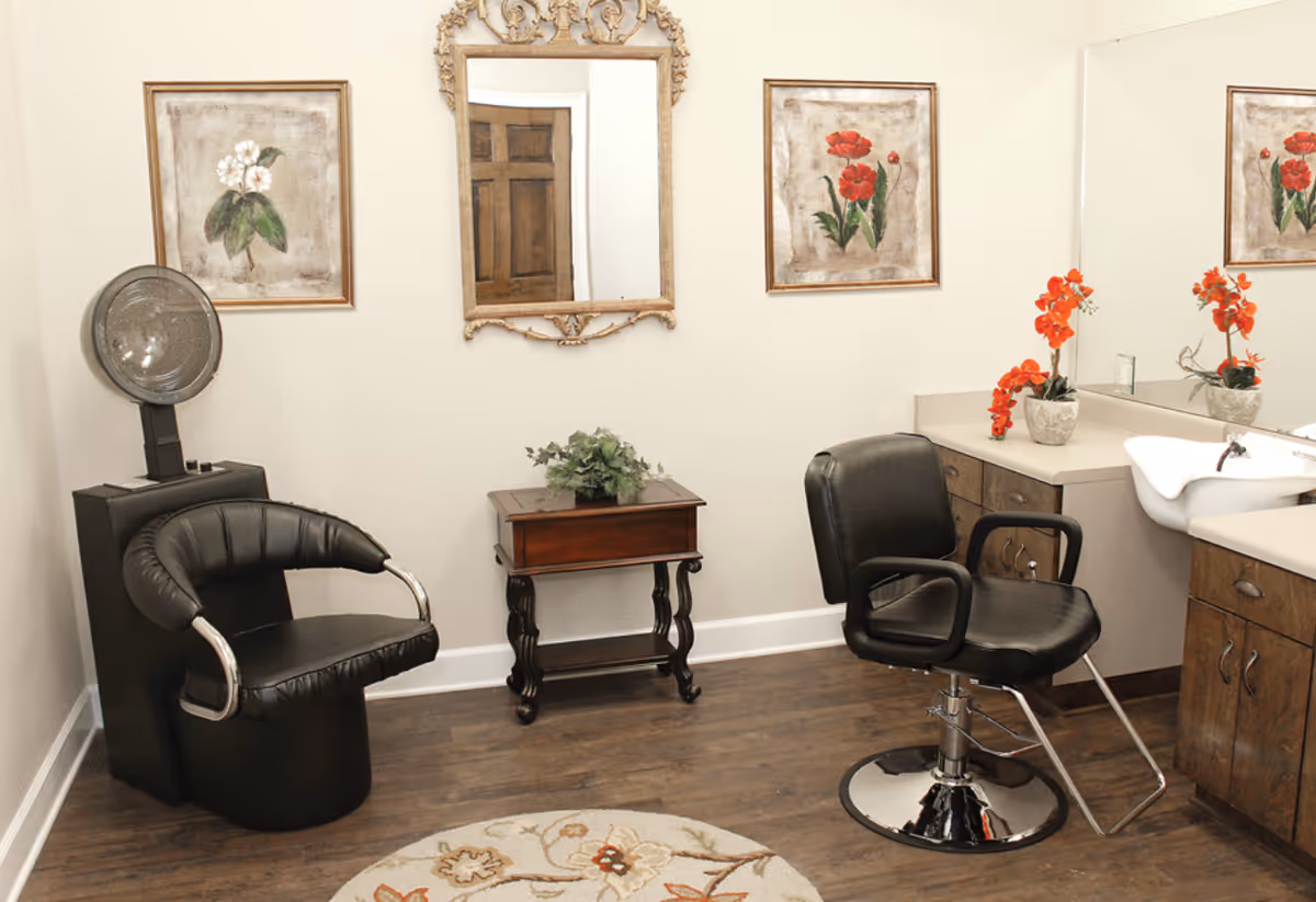 A small salon room with two black salon chairs, a hair dryer, a wooden side table with a plant, a large mirror with an ornate frame, floral paintings on the wall, and a countertop with a sink and orange flowers in a vase.