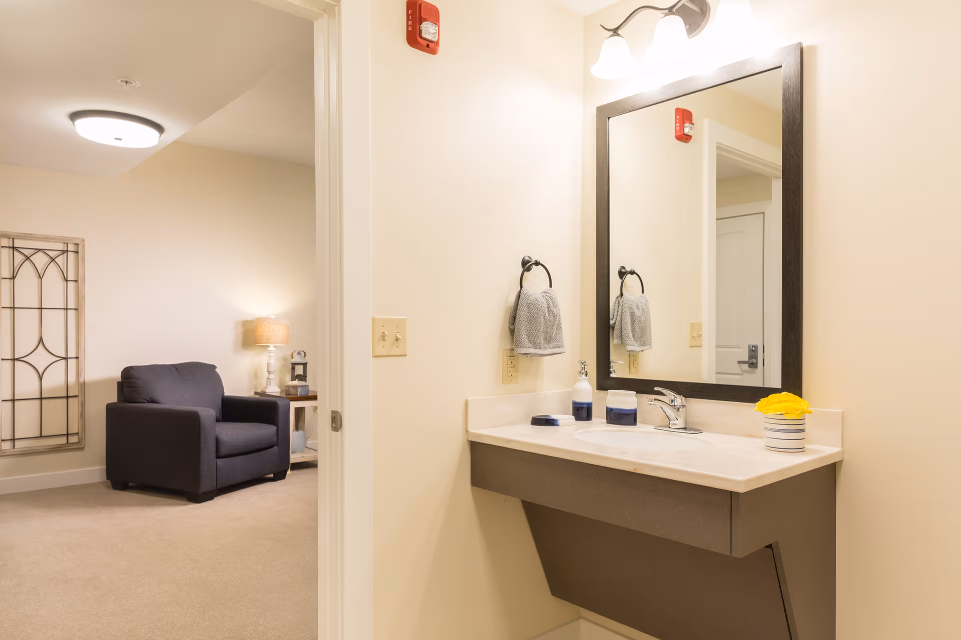 A bathroom vanity with a mirror, sink, and towel beside an open doorway leading to a sitting area with an armchair and side table.
