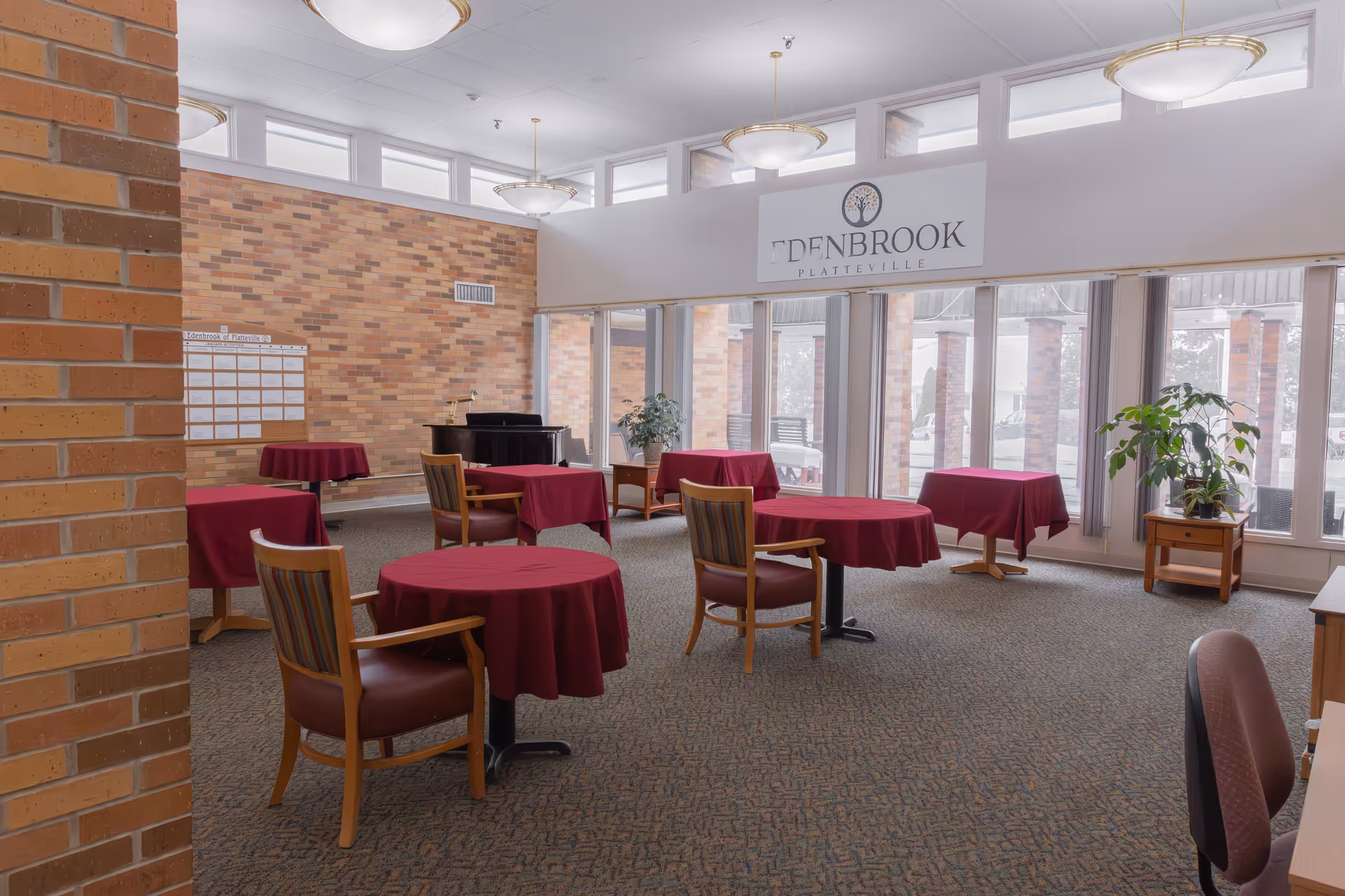 A spacious room with several round tables covered in burgundy tablecloths, each accompanied by wooden chairs with padded seats. The room has large windows letting in natural light, brick walls, and a sign that reads 'Edenbrook Platteville' above the windows. There are also some plants and a piano in the background.