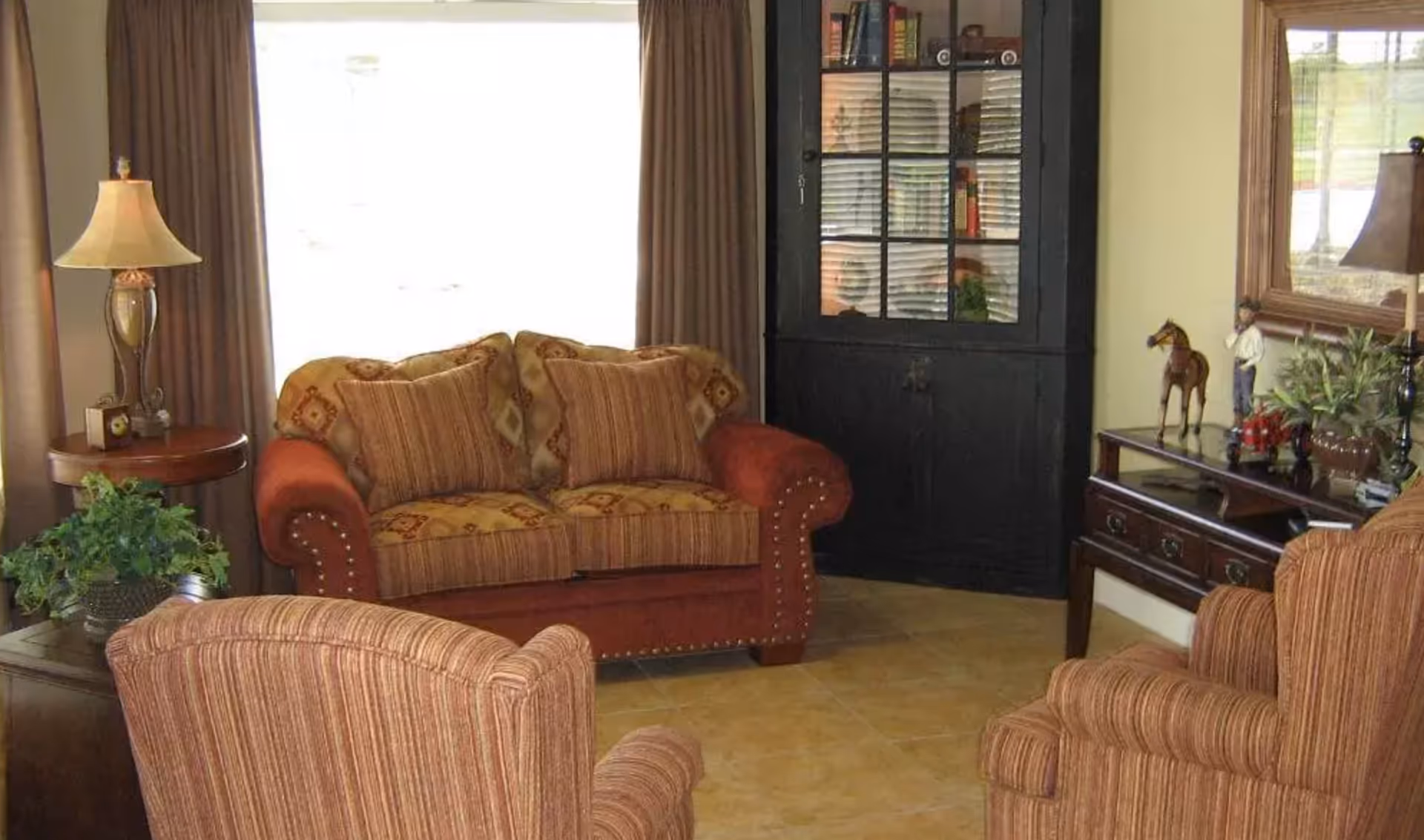 A cozy living room with a patterned loveseat and two striped armchairs arranged around a wooden side table with a lamp and a potted plant. There is a black cabinet with glass doors filled with plates and books, and a wooden console table with decorative items including a horse figurine and a lamp. A large mirror hangs above the console table.