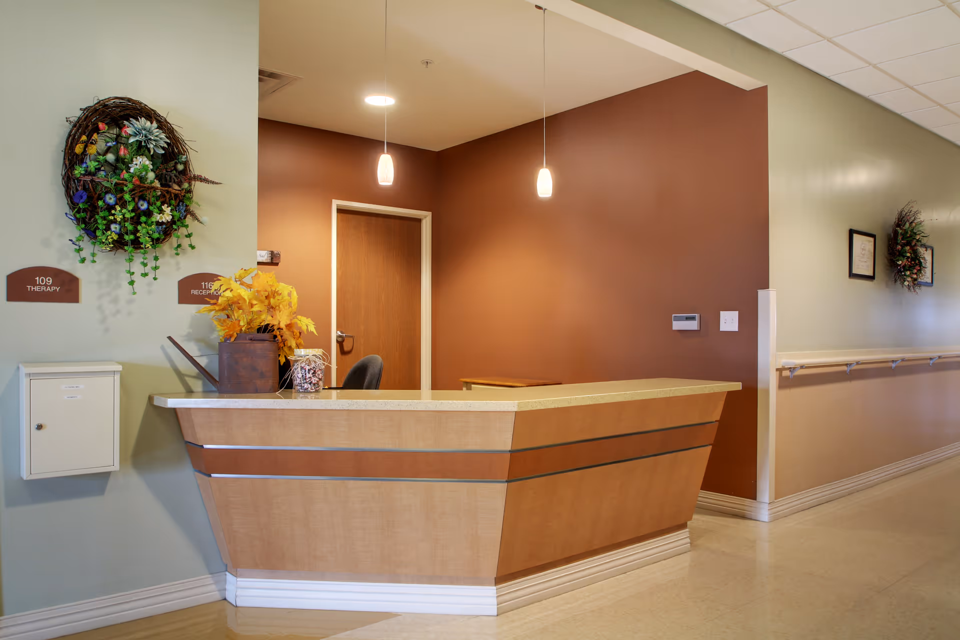 Reception desk area inside Heritage Health: Therapy & Senior Care - Hoopeston with a wooden counter, two pendant lights hanging from the ceiling, a door behind the desk, and decorative wreaths on the walls.