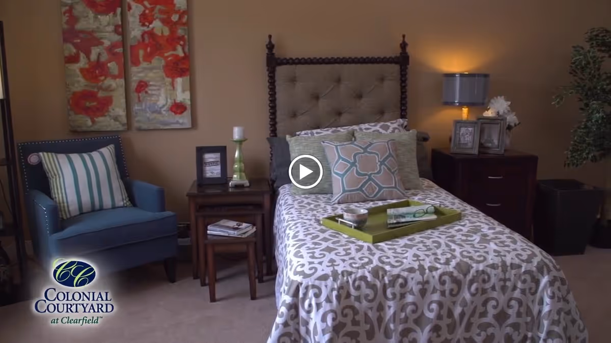 A cozy bedroom featuring a single bed with patterned bedding and multiple pillows, a green tray with a book and glasses on the bed, a blue armchair with a striped pillow, two wooden nightstands with a lamp, framed photos, a candle, and decorative items, and a potted plant in the corner. Two floral paintings hang on the wall above the armchair.