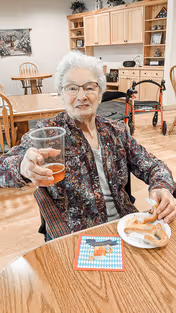An elderly woman with white hair and glasses sits at a wooden table in a dining area, holding up a glass of amber-colored liquid and smiling. On the table in front of her is a plate with pretzels and a blue and white checkered napkin. Behind her, there is a kitchen area with wooden cabinets, a walker, and another wooden table with chairs.