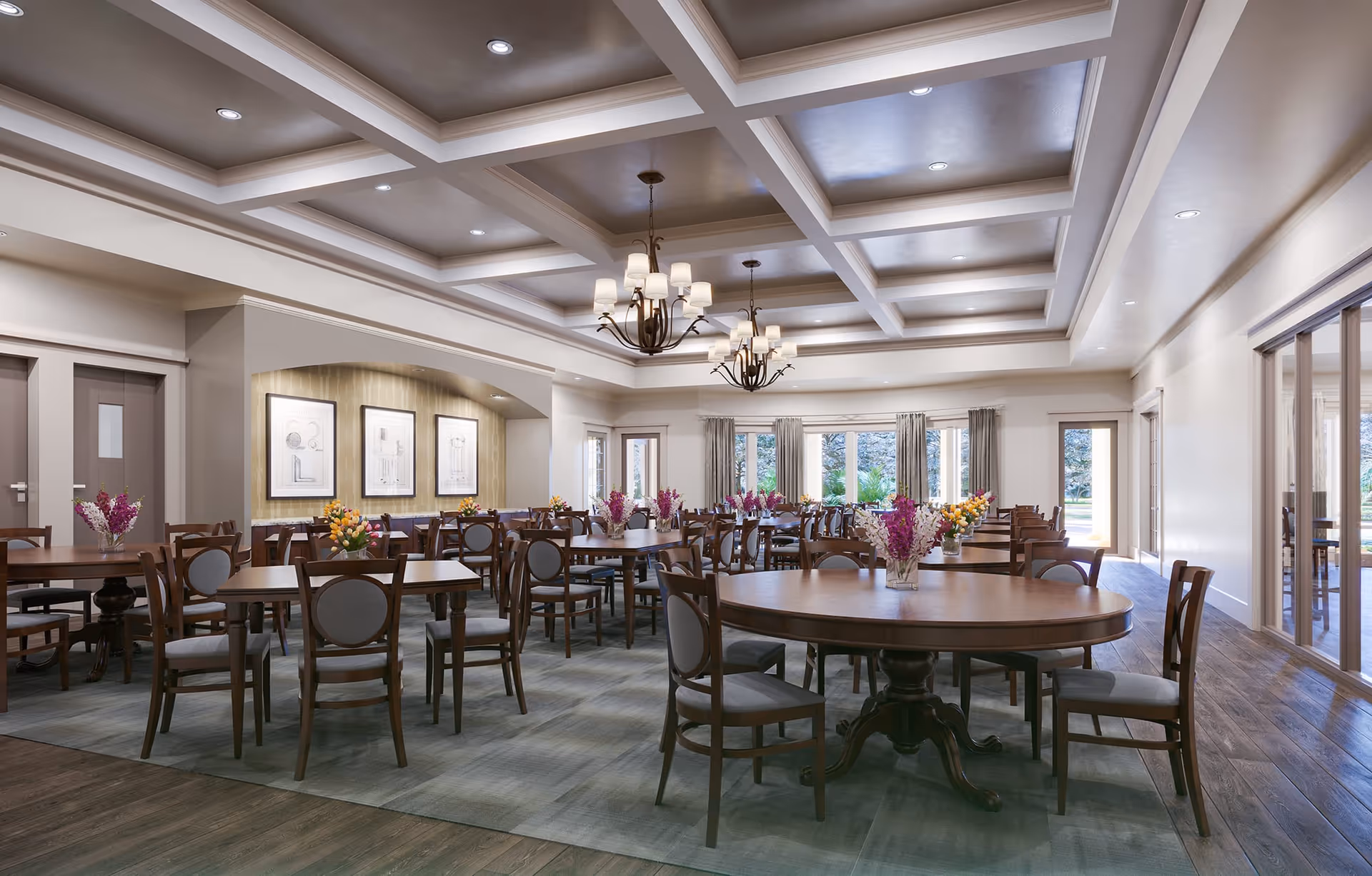 A spacious dining room with multiple wooden tables and chairs arranged neatly. Each table has a vase with colorful flowers. The room features a coffered ceiling with recessed lighting and two large chandeliers. Large windows and glass doors allow natural light to fill the room, and framed artwork is displayed on one wall.