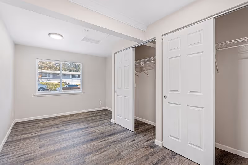 Empty room with wood flooring, a window showing an outside view of a building and cars, and two open closets with white sliding doors and hanging rods inside.