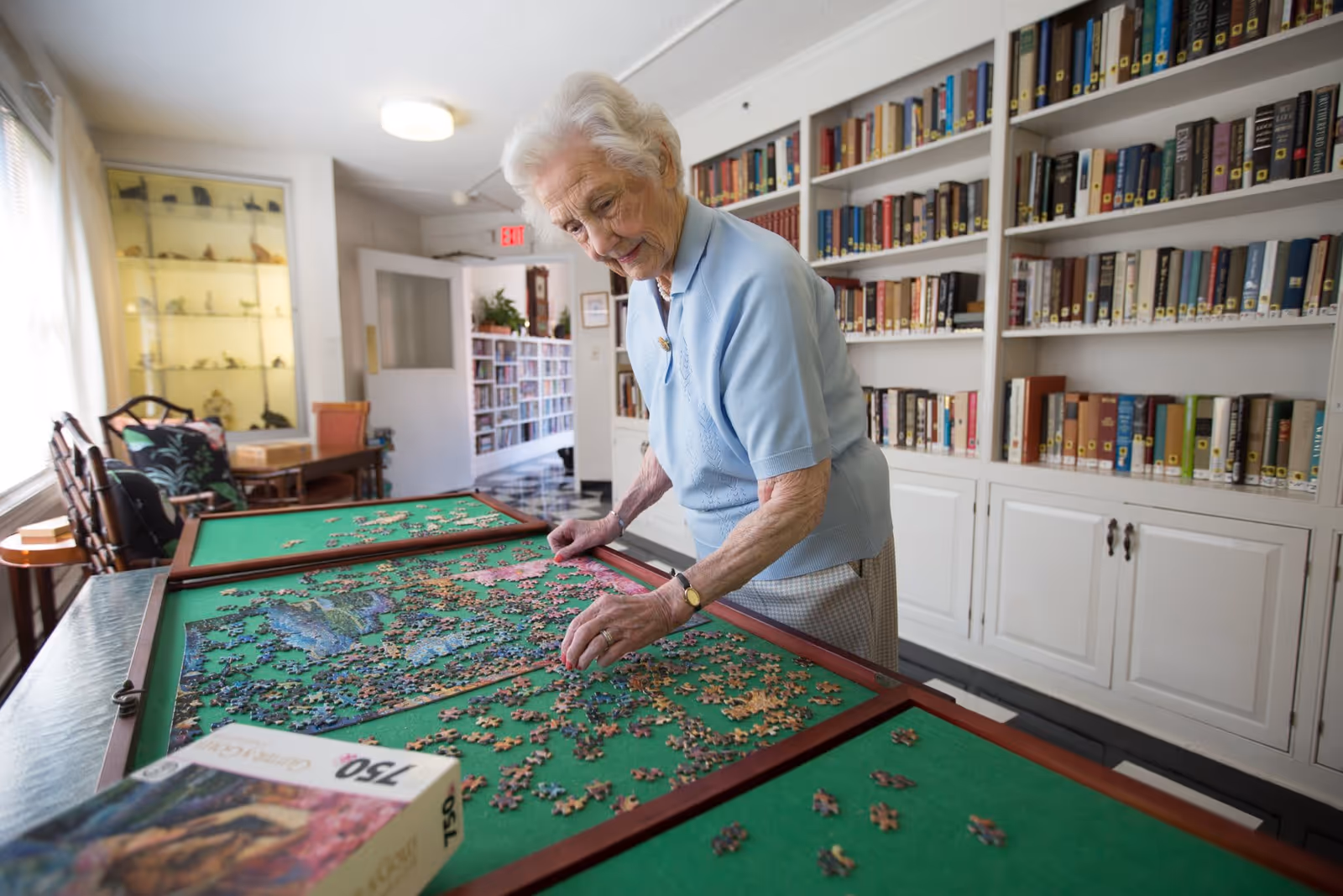 An elderly woman with white hair is working on assembling a jigsaw puzzle on a green felt table in a room with white bookshelves filled with books. The room is well-lit with natural light coming from a window on the left side. There are chairs and a display cabinet with small figurines in the background.