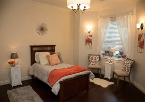 A cozy bedroom with a single wooden bed featuring white and orange bedding. The room has two white nightstands with lamps and flowers, two chairs with floral cushions near a window with white curtains, wall decorations including floral paintings and a metal wall art piece, and dark wooden flooring with two small rugs.