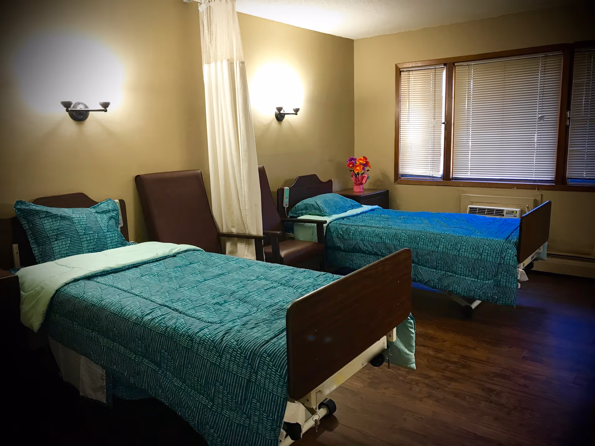 Shared senior facility bedroom with two hospital-style beds dressed in teal bedding, chairs, a privacy curtain, and a window with blinds.