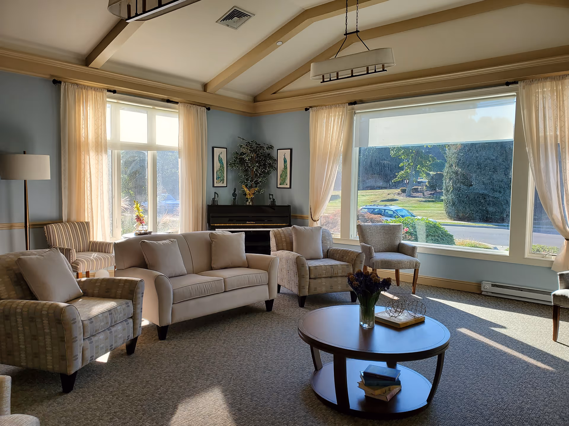 A bright and cozy living room with large windows letting in natural light. The room features a beige sofa, several armchairs, a round wooden coffee table with books and a vase of flowers, a floor lamp, and a piano against the wall. Light curtains frame the windows, and the walls are painted a soft blue with cream trim.