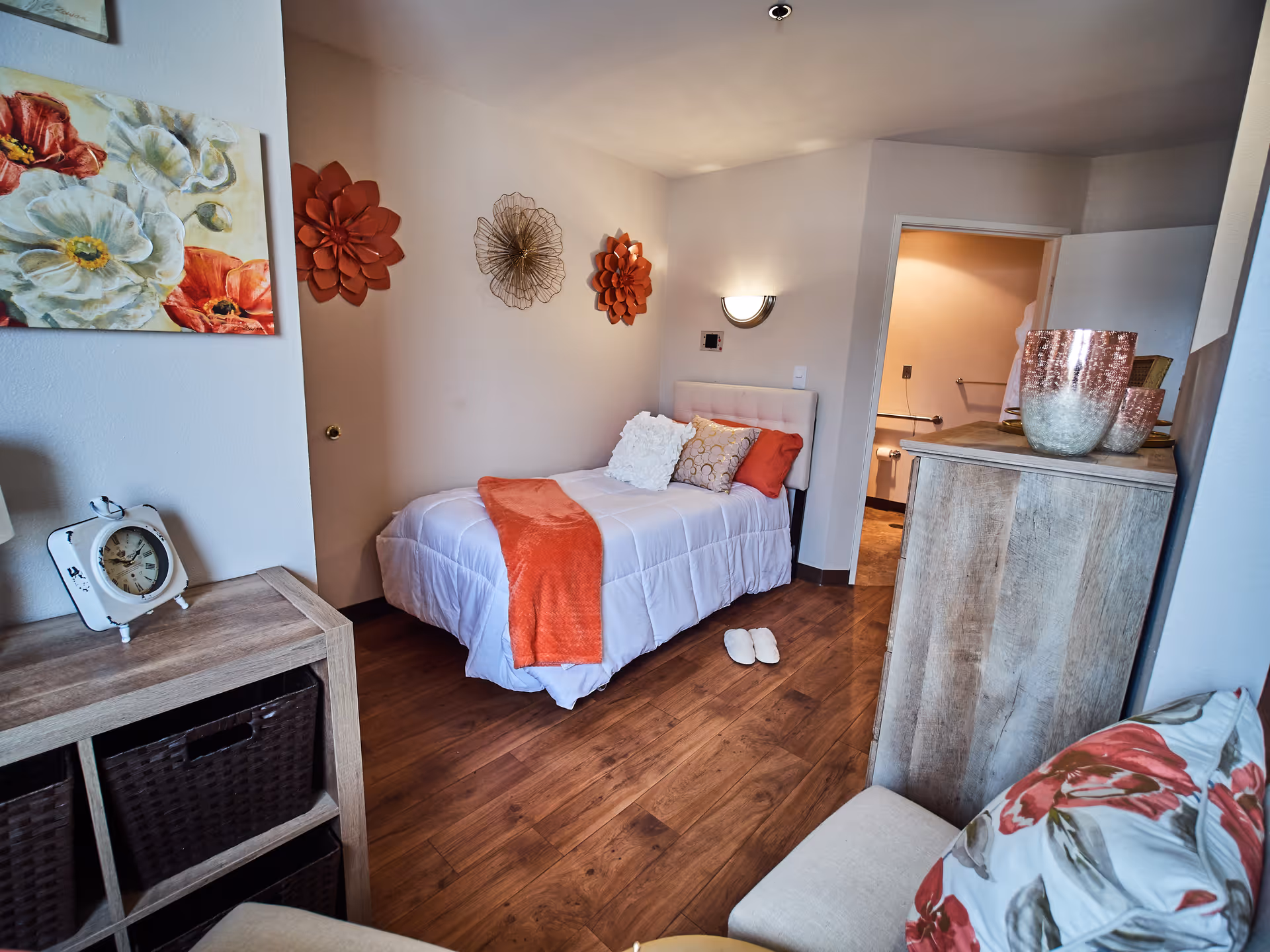 A cozy bedroom in a senior living facility featuring a single bed with white bedding and orange accents, including a throw and pillows. The room has wooden flooring, decorative wall art with floral designs, a wooden dresser with decorative vases, a small clock on a wooden shelf, and a cushioned chair with a floral pillow. An open door leads to a bathroom.
