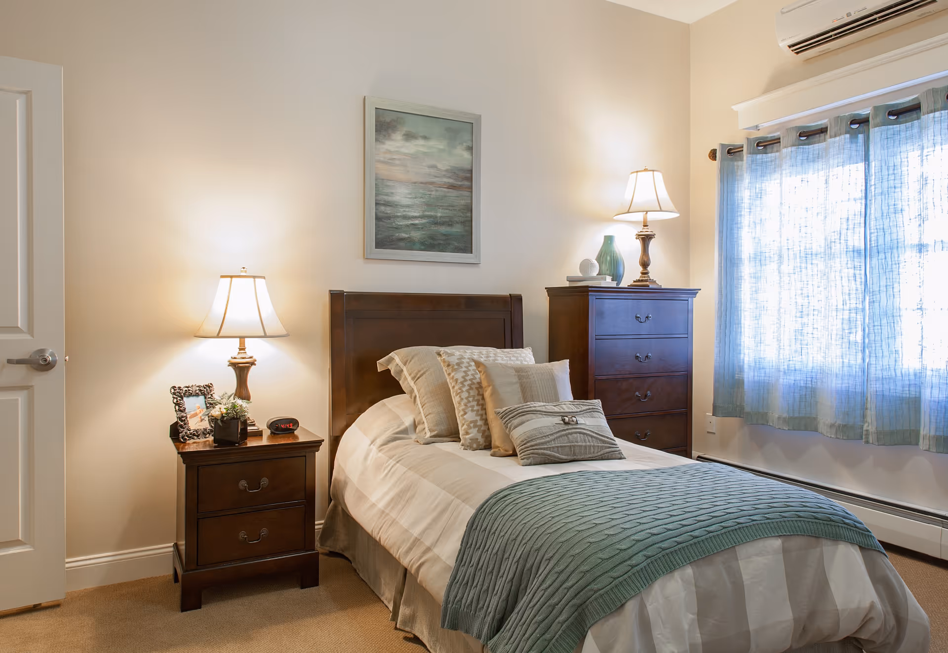 A cozy bedroom with a single bed featuring multiple pillows and a green knitted throw blanket. There is a wooden nightstand with a lamp, a framed photo, a small plant, and an alarm clock. A wooden chest of drawers with a lamp, books, and a decorative vase is next to a window with light blue curtains. The walls are light-colored, and there is a framed painting above the bed.