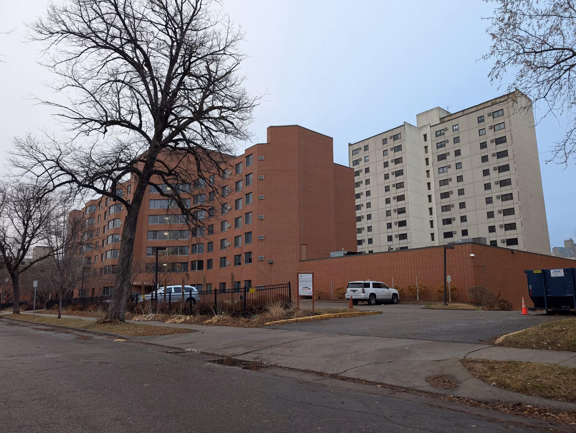 Exterior view of a multi-story brick and concrete apartment building with bare trees and parked cars in front.