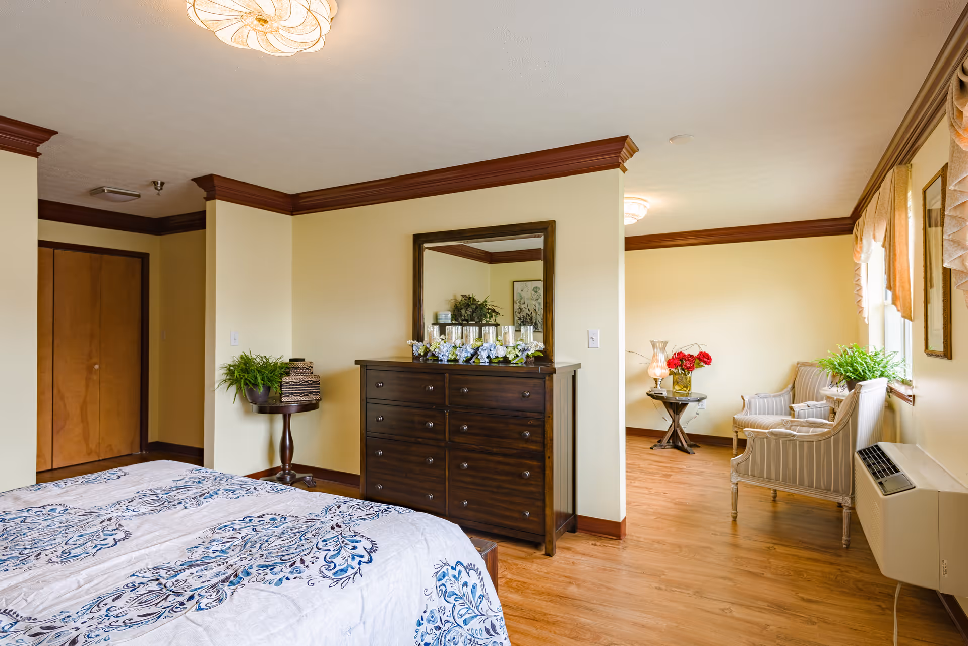 A bright bedroom in Brentwood Retirement Community featuring a bed with a patterned blue and white bedspread, a dark wooden dresser with a large mirror above it, a small round table with a plant, and a sitting area with two striped armchairs and a small table with a lamp and flowers near a window with curtains.